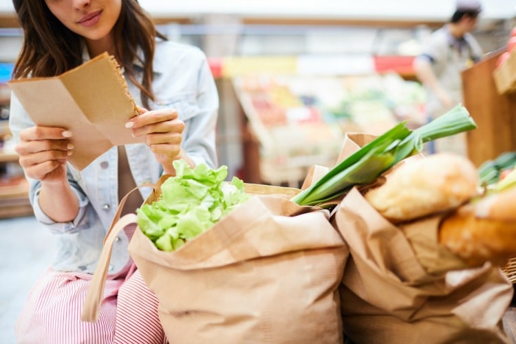 Close-up of content young woman crouching near full shopping bags and reading shopping list in organic food store